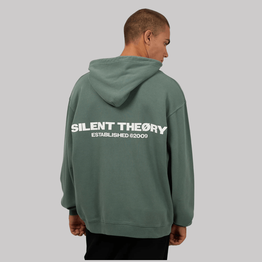 Back view of man wearing a green hoodie with a logo on a gray background. Silent Theory Essential Theory Hoodie with front and back logo print available at Stewart’s Menswear Mullumbimby.