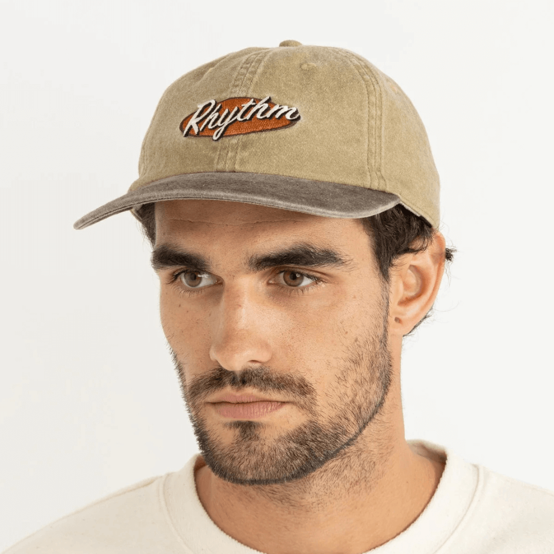 Stewart's Menswear, Mullumbimby. Rhythm Hallmark Cap. Sand coloured with embroidered Rhythm branding and darker brown coloured peak. Photo Shows model wearing Hallmark Cap, front view.