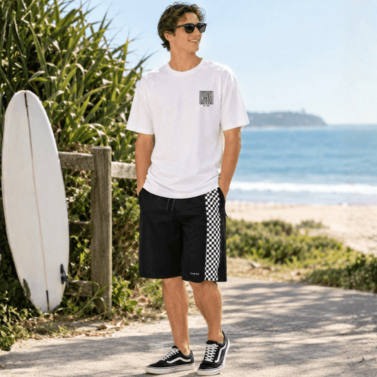 A man stands on a beach path wearing Carve WAVEY t-shirt with Carve Checkmate Boardshorts and black-and-white sneakers. A white surfboard leans against a wooden fence beside him, with the ocean and sandy beach in the background.
