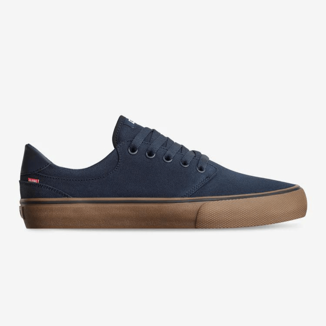 Stewarts Menswear Globe Goodstock Skate Shoe. The Globe Goodstock skate shoe in Navy/Gum is a classic Globe silhouette with a canvas upper. Lace-up, fully lined mainstay style featuring a Super V outsole for enhanced grip and board feel and a taller rubber foxing for maximum durability. Top is Navy canvas with Gum coloured sole. Photo shoes side view of shoe.