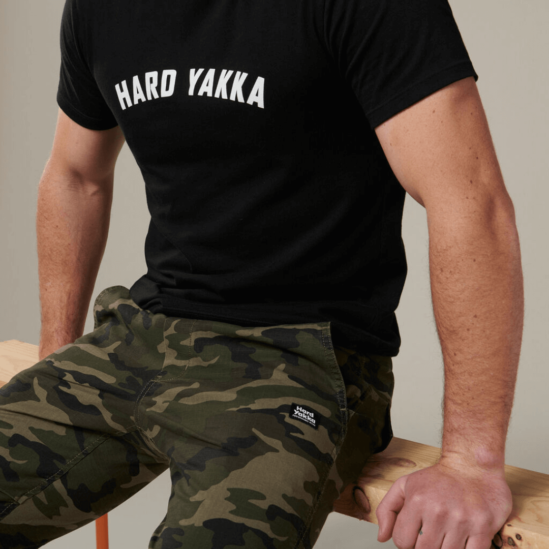 Stewarts Menswear Hard Yakka Camo Jogger Pants. These trendy Camo print jogger pants have an elasticated waistband for the perfect fit and elasticated cuff to fit comfortably over a work boot/sneaker. Photo shows a photo of seated model wearing Hard Yakka Camo Jogger pants with a black tee-shirt.