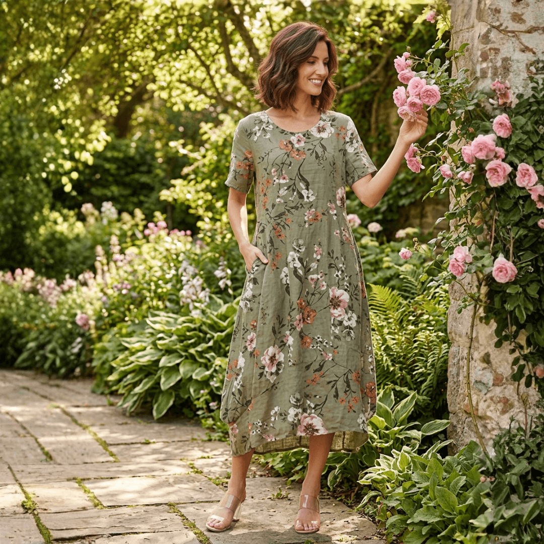 Woman wearing a khaki green floral linen midi dress, standing beside a flowering climbing rose. Harlow Lane short sleeve floral dress in khaki available from Stewart's Menswear in Mullumbimby.