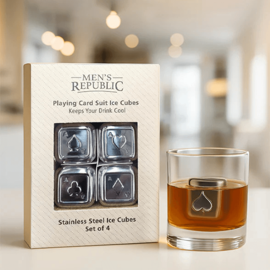 A set of four stainless steel playing card suit ice cubes in their branded Men's Republic box, shown next to a clear glass with amber liquid and one ice cube visible inside, against a softly blurred indoor background.