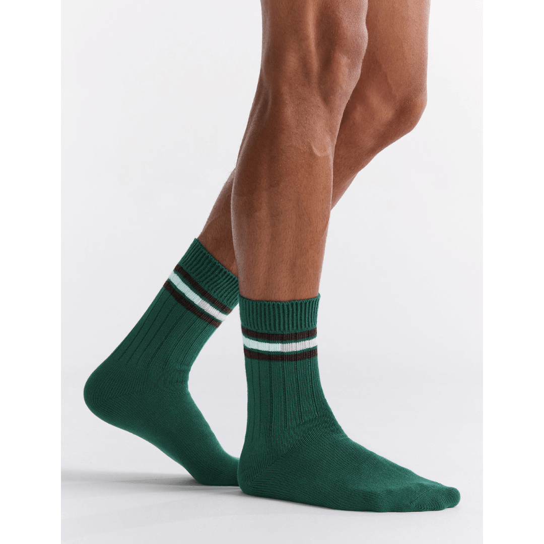 Stewarts Menswear Mullumbimby. Ambra Men's Chunky rib tennis stripe crew socks. Organic Cotton socks. Bottle green colour with three stripes at top (2 x black with 1 x white in between). Photo shows model wearing Ambra men's chunky rib tennis crew socks in evergreen.