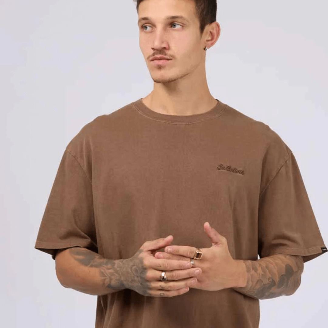 Stewarts Menswear, Mullumbimby. St Goliath Essential Script Tee. Photo shows model wearing brown coloured St Goliath Essential Script Tee with embroidered logo on left chest.