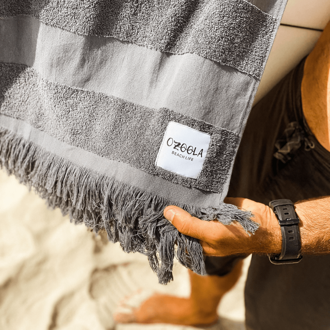 Stewarts Menswear Ozoola Pool to Beach towel.The newest addition to the OZoola Beach Towels range is the Pool to Beach towel. Perfect for endless beach days, lazy pool days, and travelling. Soft and light, this towel features a combination the very popular Turkish Cotton towels with the addition of terry stripes to keep you dry in style. Colour is Rocky (charcoal grey colour). Photo is lifestyle shot showing close up of terry stripes.