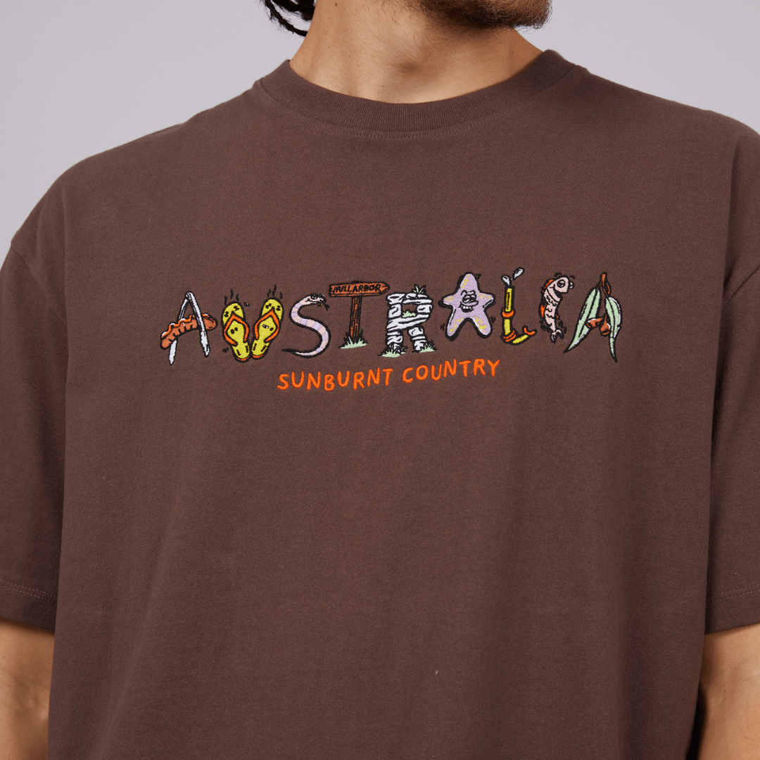 Stewart's Menswear St Goliath Australia Tee. The Australia Tee by St. Goliath is a must have holiday tee. A short sleeve tee with crew neckline made from 100% cotton. The word "Australia" is embroidered on front. Each letter is signified by something uniquely Australian including a sausage in tongs, thongs, and a snake. Colour is brown. Photo shows close up photo of embroidered Australia logo.