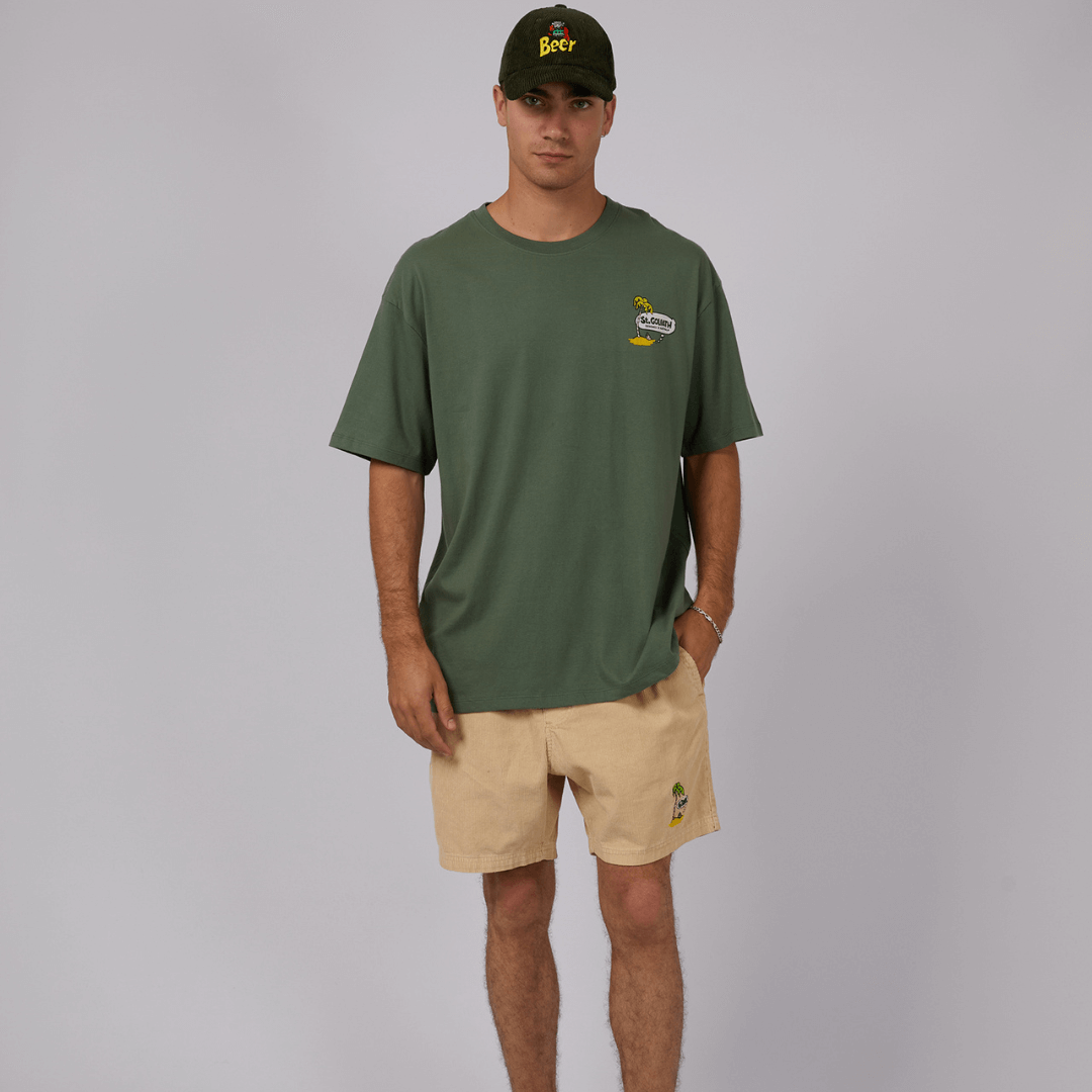 Stewarts Menswear St Goliath Souvenier Tee. The Australiana print on the back is a journey into the heart of Australia. The small print on the front makes a statement without saying a word. Easily pair with Cord Shorts and a cord cap for a relaxed look. Photo is model wearing olive coloured Souvenier t-shirt with small palm tree australiana logo on the front.
