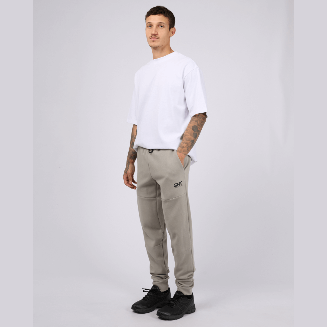 Stewarts menswear mullumbimby, St Goliath Tech Track pant. Colour is grey marle. Cuffed legs, black St Goliath logo on front side pocket. Photo shows full model front view with white t-shirt.