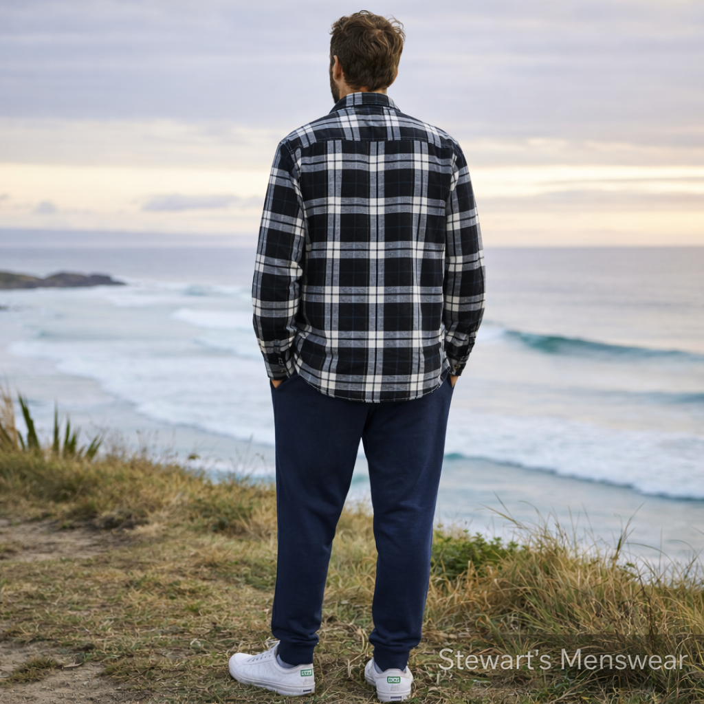 Man wearing a black and white plaid shirt and navy pants standing on a grassy area overlooking the ocean. Pilbara Collection Flannelette Shirt classic check 160gsm cotton, available at Stewart’s Menswear Mullumbimby.
