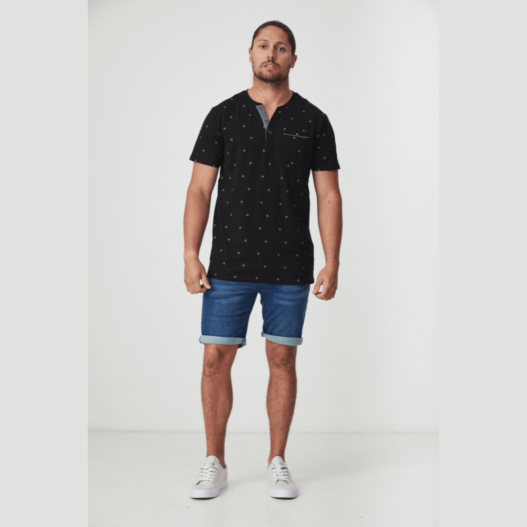 Stewarts Menswear Blackwood Apparel Arnold Henley neck tee-shirt. The Blackwood Apparel Arnold Short Sleeve Henley Tee is made from 100% cotton, combining classic comfort with modern durability. Colour is black with a tiny geometric printed all over. Chest pocket and contrast fabric in placket. Model wearing Arnold Henley tee with Perth Denim shorts.
