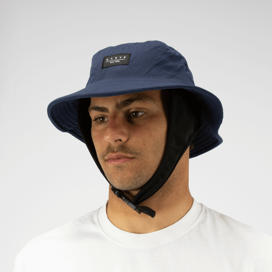 Model wearing a Navy coloured Carve Big Wave Surf Hat, available at Stewart’s Menswear Mullumbimby. Quick-dry polyester surf bucket hat with chin strap and logo badge.