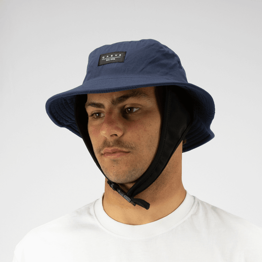 Model wearing a Navy coloured Carve Big Wave Surf Hat, available at Stewart’s Menswear Mullumbimby. Quick-dry polyester surf bucket hat with chin strap and logo badge.