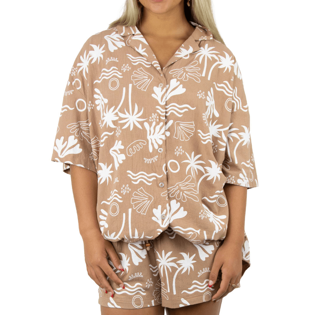 Stewart's Menswear Carve Ladies Las Palmas Short set. Ladies short and shirt matching set.  Short sleeve loose fit button through shirt with elastic waist shorts featuring all over palm/beach print and Carve flag on side seam. Colour is Mochi with white yardage print featuring beach themed abstract shapes.