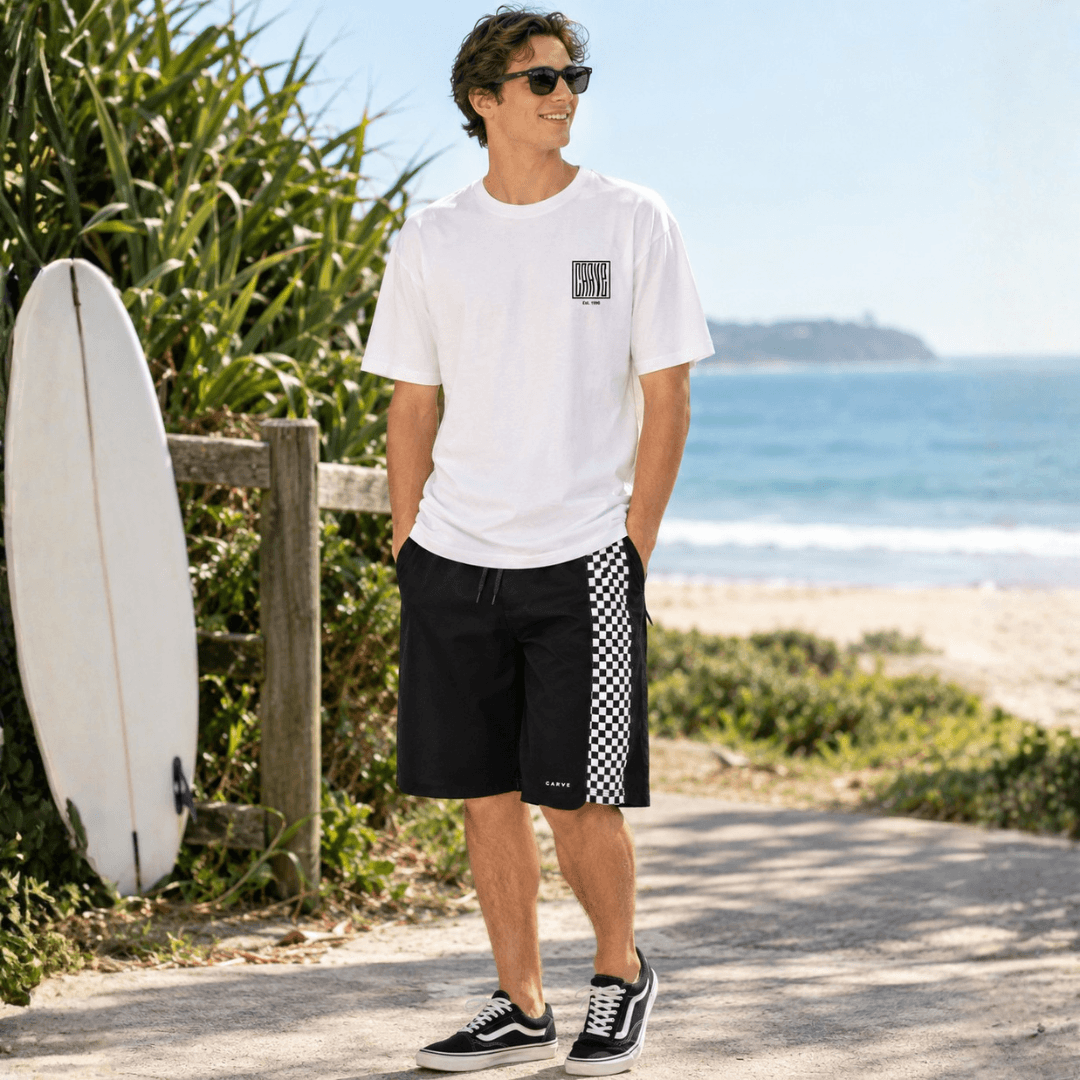 A man stands on a beach path wearing Carve WAVEY t-shirt with Carve Checkmate Boardshorts and black-and-white sneakers. A white surfboard leans against a wooden fence beside him, with the ocean and sandy beach in the background.