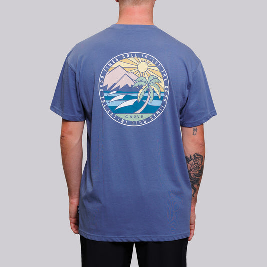 Stewarts Menswear Carve Surfwear Good Times T-shirt. This is a regular fit men's short sleeve T-shirt with small graphic chest print and large back graphic print. With sizes from S to 5XL, no-one is excluded from having up to date seasonal fashion. Model is wearing indigo coloured shirt showing back print. Mountain and waves coloured graphic in circle with words "Let the good times roll in"