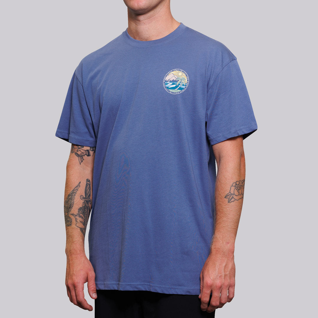 Stewarts Menswear Carve Surfwear Good Times T-shirt. This is a regular fit men's short sleeve T-shirt with small graphic chest print and large back graphic print. With sizes from S to 5XL, no-one is excluded from having up to date seasonal fashion. Model is wearing indigo coloured shirt showing front print. Mountain and waves coloured graphic in circle with words "Let the good times roll in"