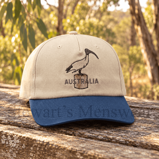 Bin Chicken Cap in stone with a blue brim, a 6-panel cotton cap with an embroidered ibis on a timber post and Australia lettering, available at Stewart’s Menswear Mullumbimby.