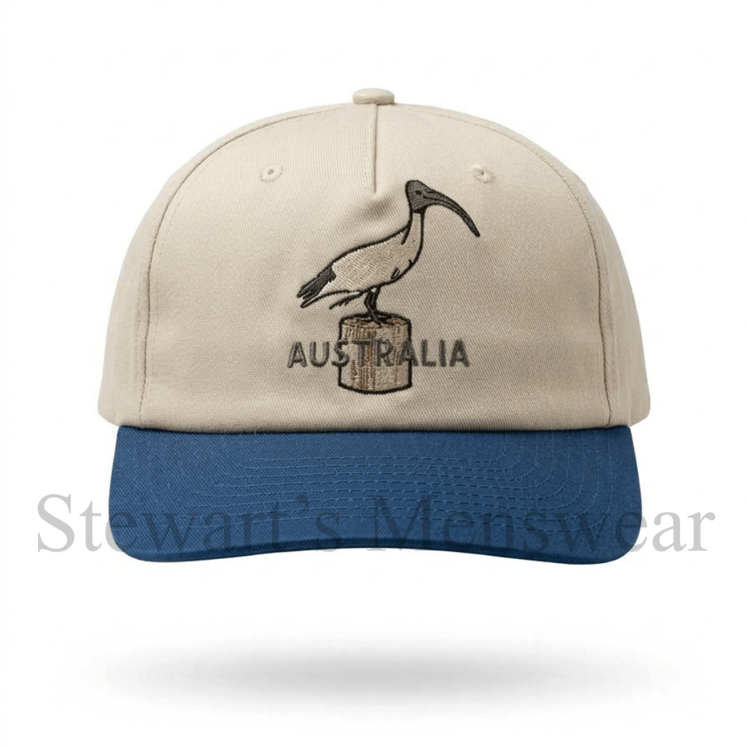 Bin Chicken Cap in stone with a blue brim, a 6-panel cotton cap with an embroidered ibis on a timber post and Australia lettering, available at Stewart’s Menswear Mullumbimby.
