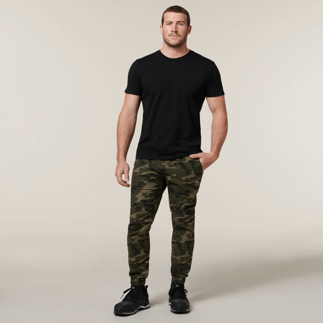 Stewarts Menswear Hard Yakka Camo Jogger Pants. These trendy Camo print jogger pants have an elasticated waistband for the perfect fit and elasticated cuff to fit comfortably over a work boot/sneaker. Photo shows full length photo of model wearing Hard Yakka Camo Jogger pants with a black t-shirt.
