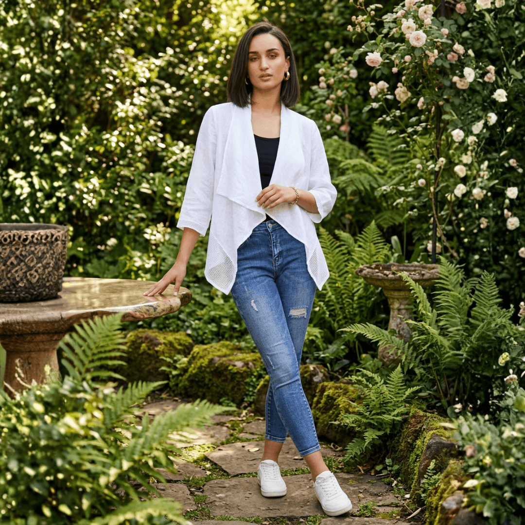Woman wearing a white waterfall linen drape top over a black cami and denim jeans, standing near a garden table. Harlow Lane mesh and linen drape top in white available from Stewart's Menswear in Mullumbimby