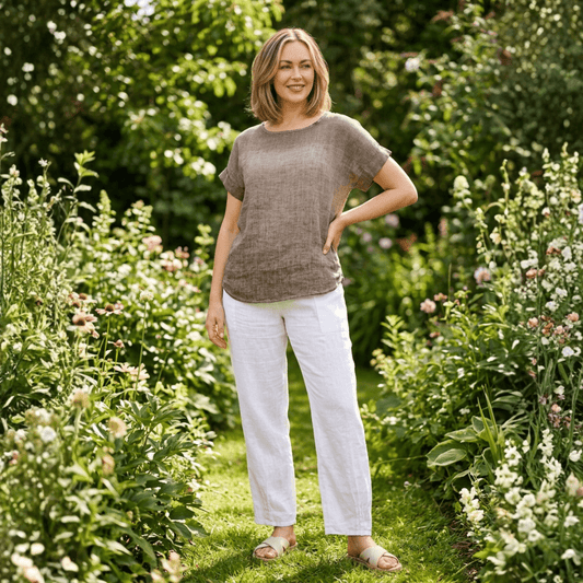 Woman wearing a chocolate brown linen top with mesh yoke detail and white linen trousers, standing among garden flowers. Harlow Lane mesh and linen top in chocolate available from Stewart's Menswear, Mullumbimby.