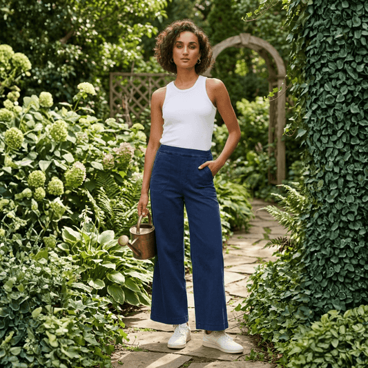 Woman wearing navy wide leg denim pants and a white tank top, standing in a garden holding a watering can. Harlow Lane wide leg denim pants available at Stewarts Menswear, Mullumbimby
