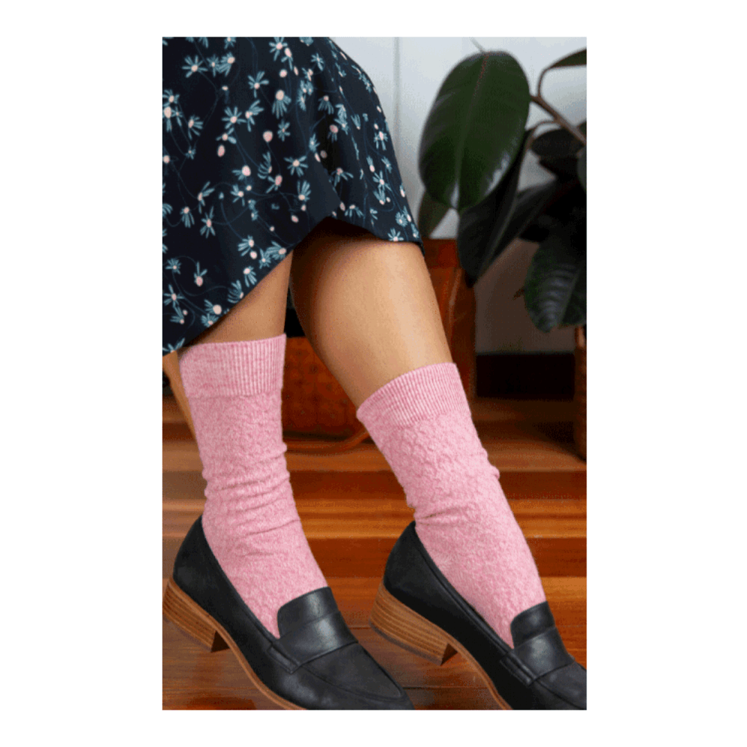 A person wearing light pink ribbed crew socks with black leather loafers, shown from the knees down in an indoor setting with a wooden floor and a potted plant in the background.