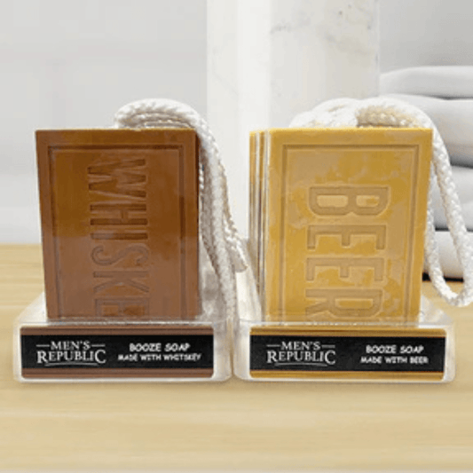 Two rectangular booze soap bars displayed on clear stands with white rope handles. The left soap is brown and embossed with 'WHISKEY,' and the right soap is tan/golden and embossed with 'BEER.