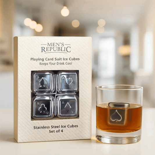 A set of four stainless steel playing card suit ice cubes in their branded Men's Republic box, shown next to a clear glass with amber liquid and one ice cube visible inside, against a softly blurred indoor background.