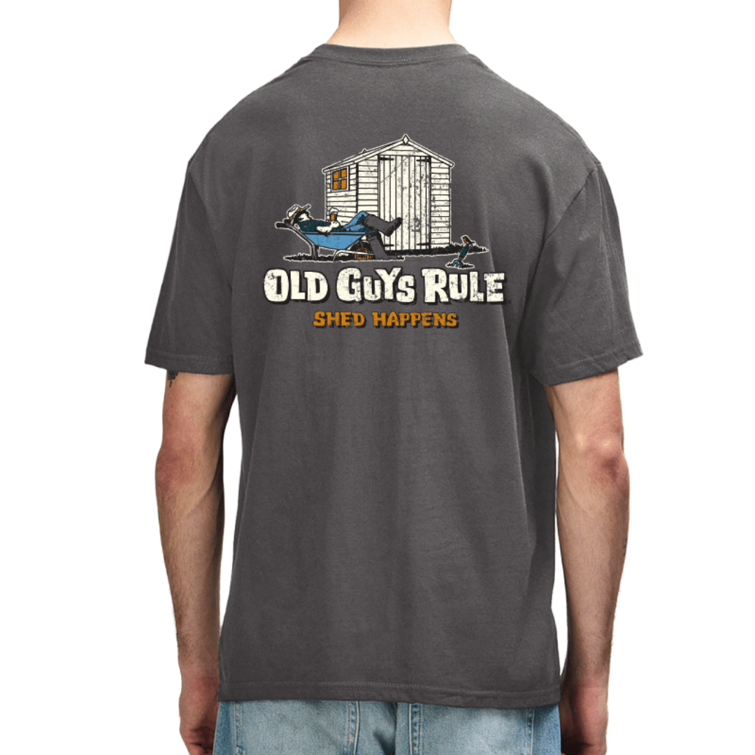 A man wearing a dark gray t-shirt, viewed from the back. The shirt features a graphic of a white shed with a man resting in front of it, with the text 'OLD GUYS RULE' and 'SHED HAPPENS' printed below.