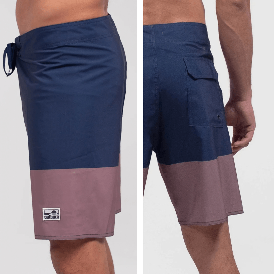 A split view of a man wearing color-block board shorts, showing the side and back pockets. The shorts feature a navy blue upper half and a dusty mauve-pink lower half, with a small black and white 'outback' logo patch on the side.