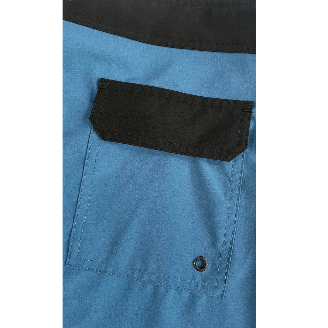 Close-up of a blue garment with a black pocket flap and black trim, showing the fabric texture and construction details.
