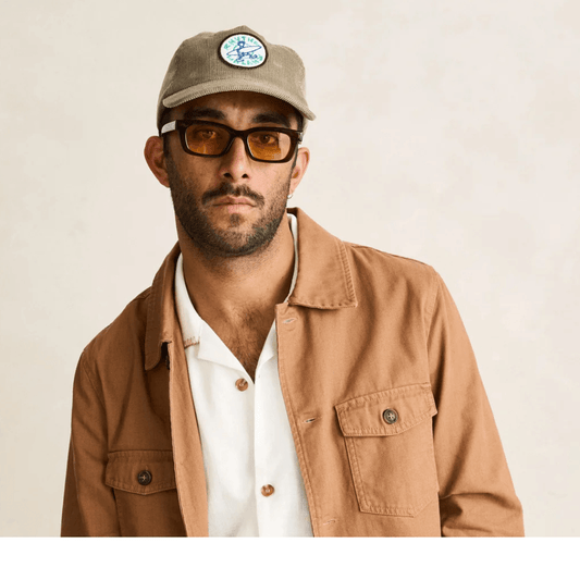 Man wearing a stone coloured corduroy baseball cap with a circular embroidered patch on the front featuring a blue and green surfer graphic. Rhythm Jaunt Cord cap available from Stewart's Menswear in Mullumbimby