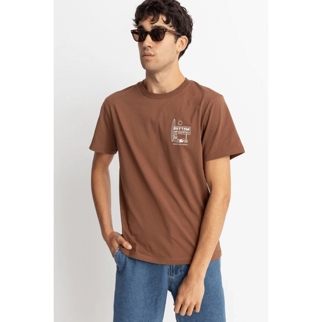 Stewarts Menswear Rhythm Livin Lull T-shirt. Colour is chocolate. Image shows front of tee-shirt with small chest print of man lying on the ground leaning on an upright surfboard with a sign above which reads: Rhythm Surf Essentials.