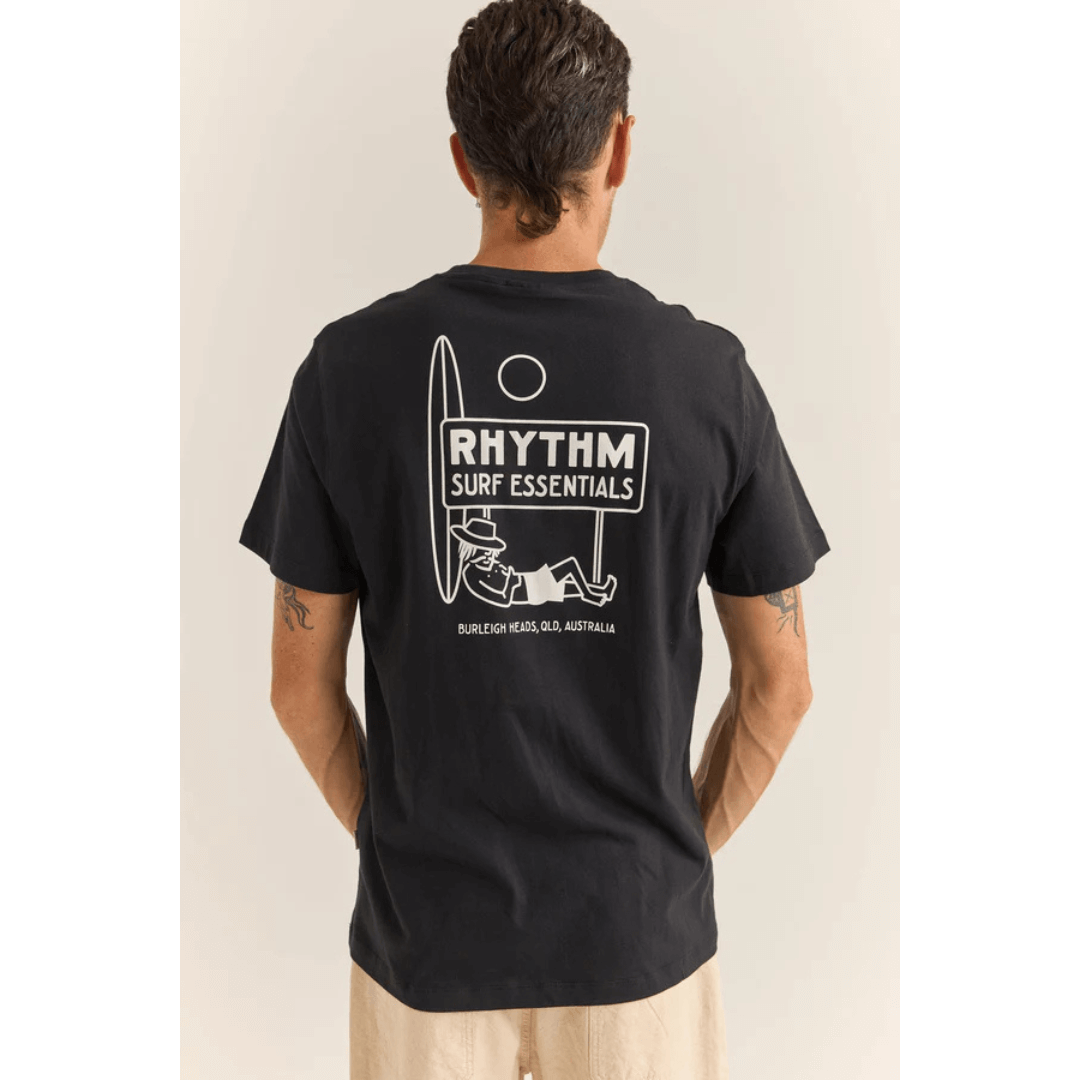 Stewarts Menswear Rhythm Livin Siesta T-shirt. Colour is black. Photo shows model wearing Rhythm Siesta Tee, back view. Large screenprint graphic on back of surfer leaning on a surfboard and lying under a sign which reads "Rhythm Surf Essentials."