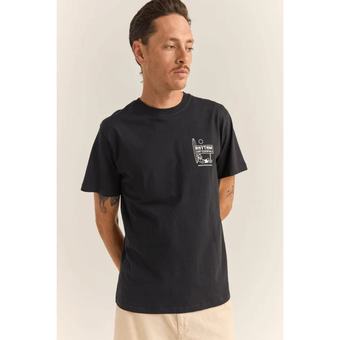 Stewarts Menswear Rhythm Livin Siesta T-shirt. Colour is black. Photo shows model wearing Rhythm Siesta Tee, front view. screenprint graphic on left chest of surfer leaning on a surfboard and lying under a sign which reads "Rhythm Surf Essentials."