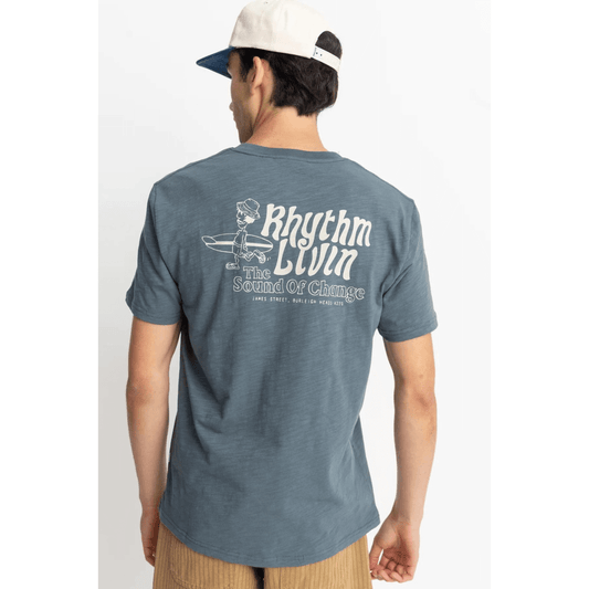 Stewarts Menswear Rhythm Slub T-shirt. Colour is Slate Blue. Large surfer carrying board print on back with text "Rhythm Livin The Sound of Change." Photo shows model wearing Rhythm Slub Tee Shirt, back view.