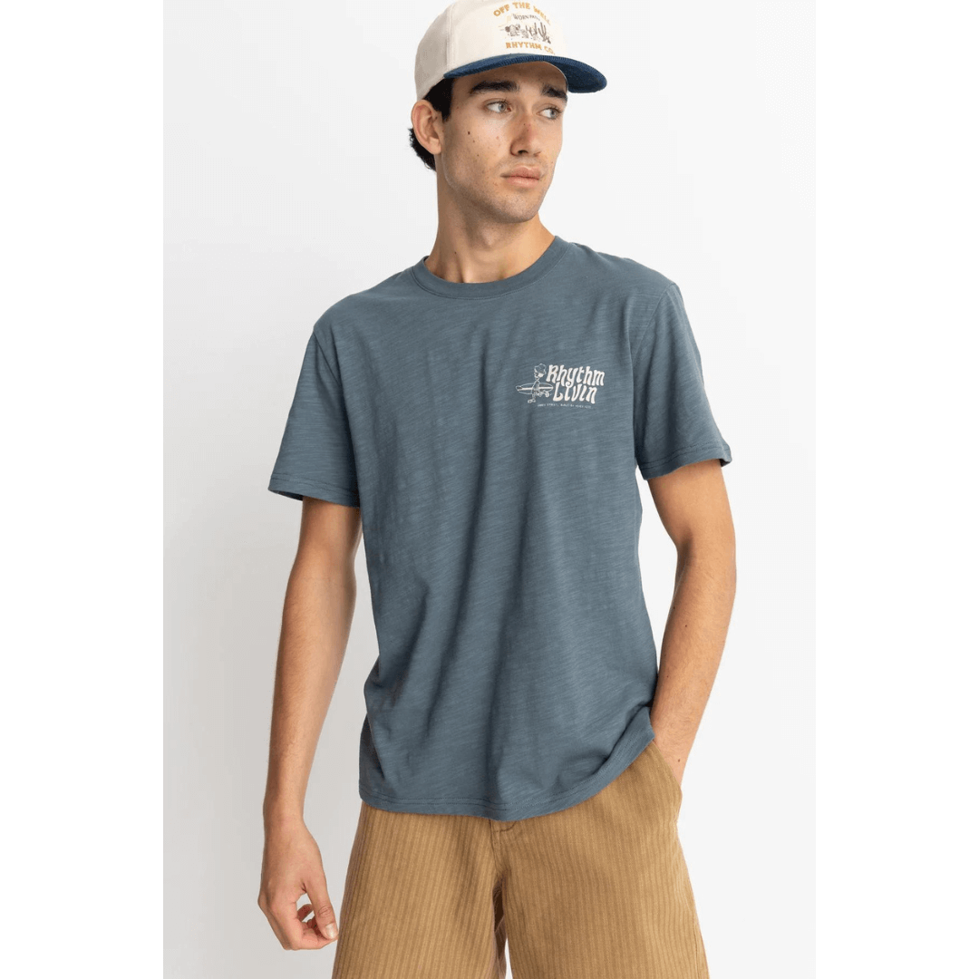 Stewarts Menswear Rhythm Slub T-shirt. Colour is Slate Blue. Small Left chest surfer carrying board print  with text "Rhythm Livin ." Photo shows model wearing Rhythm Slub Tee Shirt, front view.