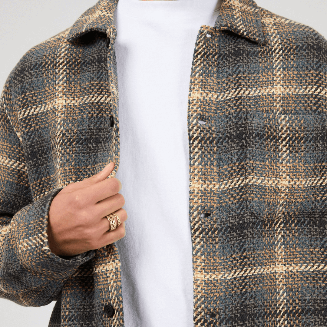 Close up view of model wearing a plaid shirt over a white tee-shirt. Silent Theory Stature Overshirt Brown in classic check with chest pockets, available at Stewart’s Menswear Mullumbimby.