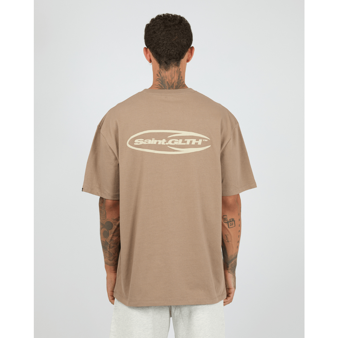 Model wearing taupe St. Goliath Omega Tee with front logo print and bold back puff-print branding, styled with grey trackpants, back view.