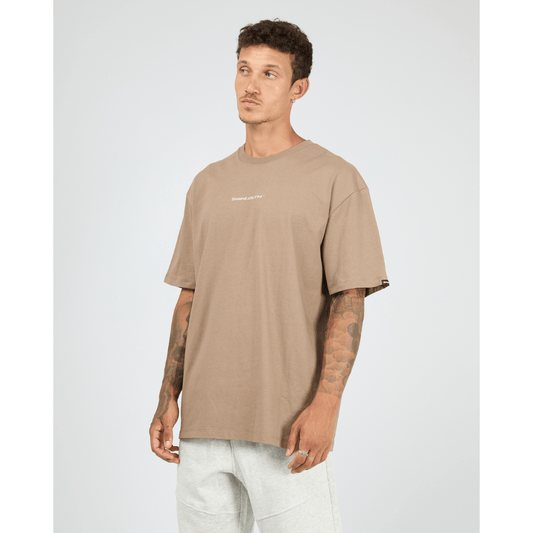 Model wearing taupe St. Goliath Omega Tee with front logo print and bold back puff-print branding, styled with grey trackpants, front view