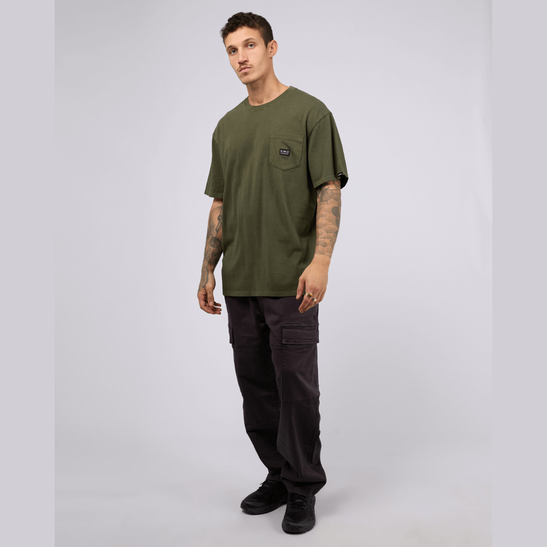 Stewarts Menswear, Mullumbimby. St Goliath pocket tee. Colour is khaki featuring a chest pocket with woven badge. Photo shows model wearing khaki coloured St Goliath pocket tee with charcoal coloured cargo pants.