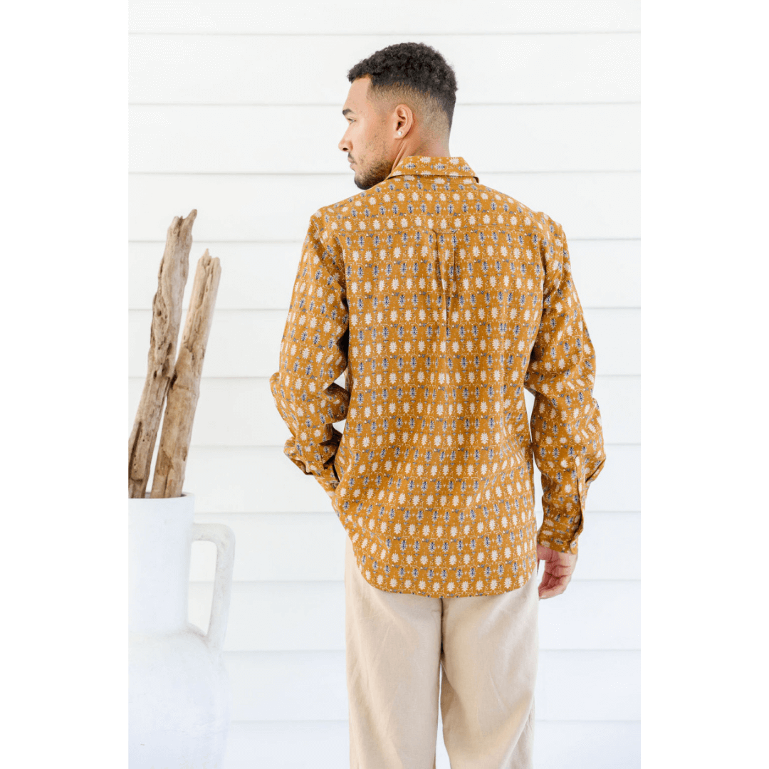 Male model is wearing a Hemp/cotton blend men's long sleeve shirt. Mustard colour with paisley like print all over. Print colours are navy, skyblue and white. Back view.