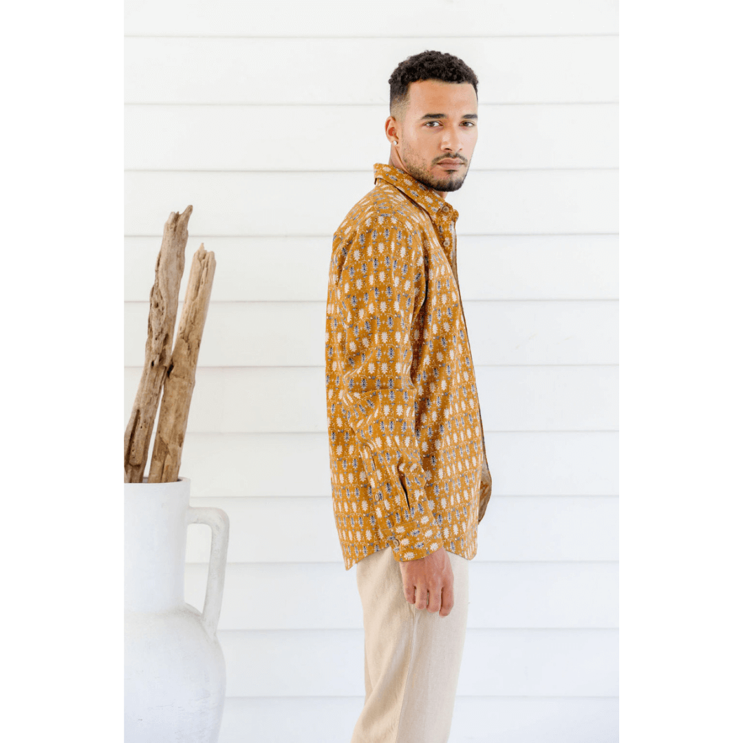 Male model is wearing a Hemp/cotton blend men's long sleeve shirt. Mustard colour with paisley like print all over. Print colours are navy, skyblue and white. Side view.